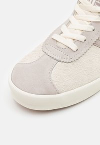 Baskets gris clair en daim et tissu texturé. Elles comportent des lacets blancs, un bout arrondi et un détail de logo subtil sur le côté.