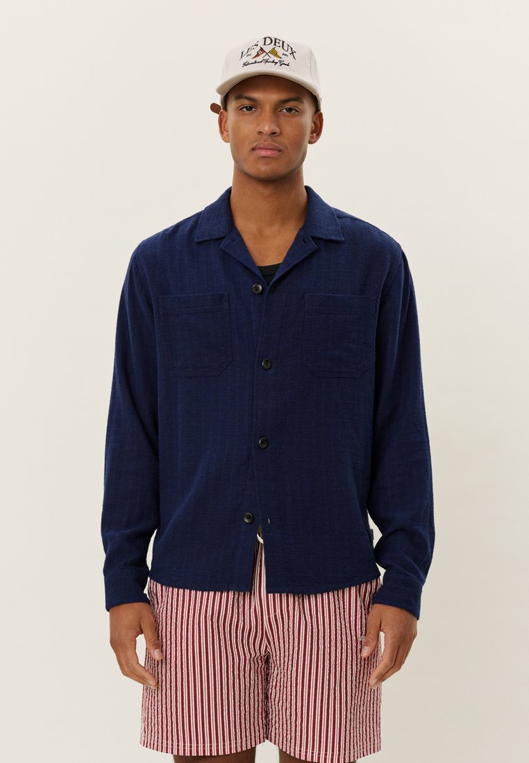 Marineblaues, knopfbares Hemd mit zwei Brusttaschen, aus leichtem Stoff und mit einer strukturierten Oberflächenbeschaffenheit. Getragen mit rot-weißen gestreiften Shorts.