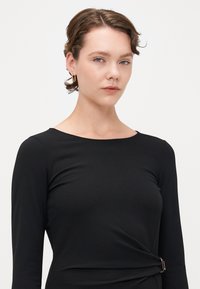 Melna kleita ar garām piedurknēm un apaļu dekoltē, ar pagrieztu detaļu jostasvietā un zelta aksesuāriem. Gluda, teksturēta auduma.