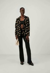 Μαύρο σακάκι με λουλουδάτο μοτίβο πάνω από ένα μαύρο κοντό τοπάκι, συνδυασμένο με μαύρα φαρδιά παντελόνια και παπούτσια χωρίς φτέρνα. Κρατάει μια ανοιχτή μπεζ τσάντα.