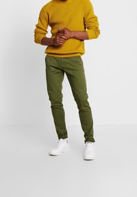 Olivgröna chinos med slim fit, med sidfickor och uppvikta benslut, kombinerade med en texturerad senapsgul tröja och vita sneakers.