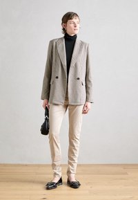 Kavaj i beige och brunt, svart polotröja, beige byxor, svarta ballerinas med rosetter och en liten svart handväska. Mot en ljus bakgrund.