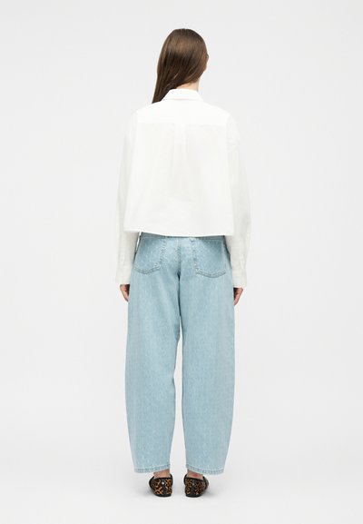 Γυναίκα που στέκεται με την πλάτη στραμμένη, φοράει λευκό κοντό πουκάμισο με μακριά μανίκια, ανοιχτό μπλε τζιν με φαρδιά πόδια και παπούτσια slip-on με λεοπάρ σχέδιο.