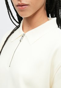 Λευκό τοπ με γιακά και φερμουάρ, με λεία υφή, διαθέτοντας ασημένιο στόμιο φερμουάρ και άνετη εφαρμογή. Κοντινή λήψη που αναδεικνύει τον γιακά και το ύφασμα.