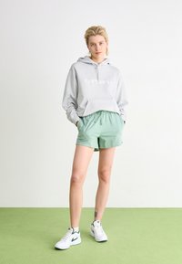 Sivi dukserica s "Carhartt" logotipom, u kombinaciji s svijetloplavim kratkim hlačama. Model nosi bijele tenisice s zelenim detaljima. Jednostavan dizajn i opušten kroj.