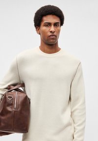 Brūna ādas plecu soma ar rāvējslēdzēja kabatu priekšpusē un īsu rokturi. Modelis valkā krēmkrāsas džemperi ar apaļu kakla izgriezumu un teksturētām piedurkņu šuvēm.