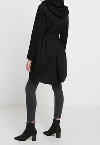 Musta hupparivelcoat, jossa on vyöllä varustettu vyötärö ja liehuvat takasaumat. Yllään tiukat farkut ja nilkkapituiset, leveäkorkoiset saappaat.