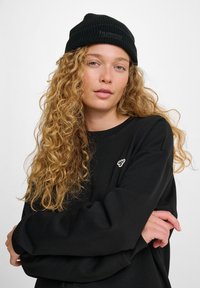 Donna con lunghi capelli ricci biondi, che indossa un berretto a maglia nero e una felpa nera, con le braccia incrociate contro uno sfondo semplice.