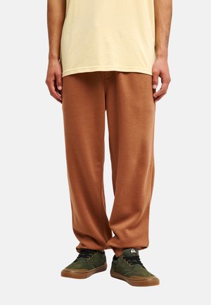 Mann trägt lockere braune Hose, grüne Sneaker mit braunen Sohlen und ein hellgelbes T-Shirt, steht vor einem einfarbigen Hintergrund.