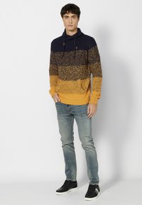 Sudadera de punto en azul marino, mostaza y gris moteado. Tiene un corte relajado, puños acanalados y un cuello con cordón. Combinada con denim claro y zapatillas negras.