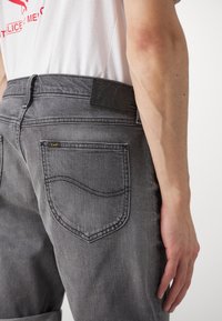 Gros plan d'une personne portant un jean gris Lee et un t-shirt blanc, montrant la poche arrière et le bras avec des veines visibles et des détails de la peau.