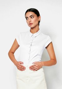 Weißes Polo-Shirt mit kurzen Ärmeln, Knopfleiste und klassischem Kragen, kombiniert mit einem cremefarbenen Rock. Glatte Textur, figurbetontes Design.