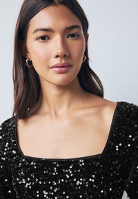 Haut à sequins noirs avec un décolleté carré et des manches longues, présentant une texture lisse et des sequins brillants, agrémenté d'une bordure noire.