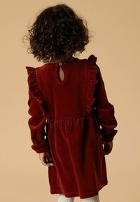 Rotes Samtkleid mit Rüschenschultern und einem Schlüssel-ausschnitt am Rücken. Lange Ärmel, geraffte Taille und plissierter Rock. Weiße Rippstrumpfhosen.