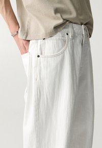 Jean en denim blanc avec une texture légèrement délavée, présentant des détails cousus en crème clair et des rivets noirs sur les poches avant.