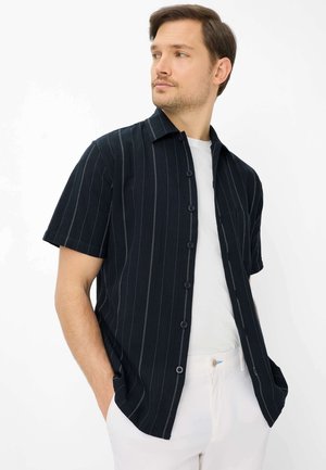 Mann trägt ein schwarzes, kurzärmliges Hemd mit dünnen weißen vertikalen Streifen über einem weißen T-Shirt und weißen Hosen, Hände in den Taschen.