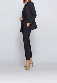 Costume noir sur mesure avec un blazer ajusté et un pantalon slim. Le tissu semble lisse, assorti à des mules beiges à talons hauts.
