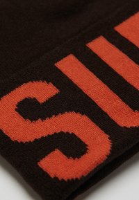 Bruine stof met een gestructureerd oppervlak, voorzien van grote oranje breiletters die "SUP" spellen. Het product is een close-up van een textielitem.
