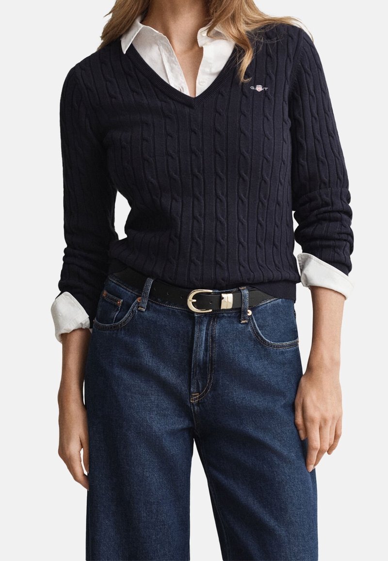 Femme portant un pull en maille torsadée sombre à col en V par-dessus une chemise blanche à col, rentrée dans un jean bleu taille haute avec une ceinture noire.