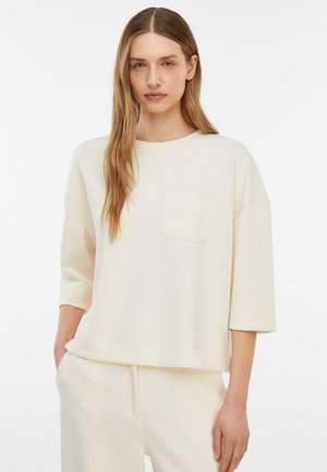 Młoda kobieta stoi z ręką w kieszeni, ubrana w luźną, kremowo-białą bluzkę z kieszenią na piersi oraz dopasowane spodnie na tle jednolitego białego tła.