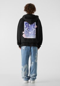 Svart hettegenser med oversized passform, med grafisk tekst "SLAY OR PLAY" i lilla og rosa, kombinert med lyseblå jeans med hvite blomsterprikker.