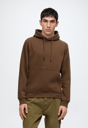 Jeune homme avec des cheveux courts et foncés portant un sweat à capuche marron et un pantalon vert olive, debout devant un fond blanc uni.