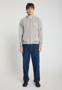 Veste à capuche zippée gris clair avec cordons de serrage, poches avant ; associée à un jean large bleu foncé et des baskets noires. Texture de tissu lisse.