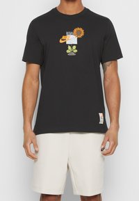 Man som bär en svart Nike-t-shirt med en solrosgrafik och vita shorts med en liten svart Nike-logga på vänster ben.
