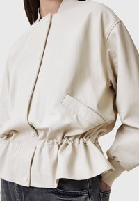Beige læderjakke med en tætsiddende talje og samlet kant, der har en knaplukning og en struktureret frontklap. Inkluderer side pocket.
