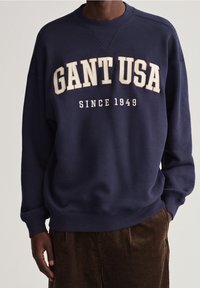 Námořnická modrá bavlněná mikina s bíle vyšitým textem "GANT USA" a "SINCE 1949", s klasickým kulatým výstřihem a žebrovanými manžetami.