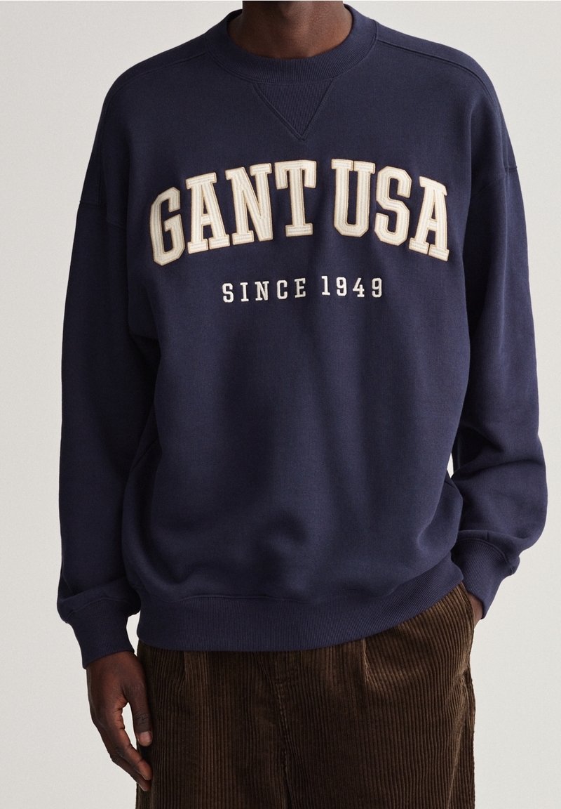 Námořnická modrá bavlněná mikina s bíle vyšitým textem "GANT USA" a "SINCE 1949", s klasickým kulatým výstřihem a žebrovanými manžetami.