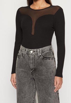 Musta pitkähihainen bodysuit, jossa on läpinäkyvä verkkoyläosa ja peittävä vartalokappale, yhdistetty korkeavyötäröisiin harmaisiin rentoihin farkkuihin.