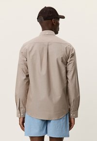 Ελαφρύ μπεζ πουκάμισο με μακριά μανίκια, πιέτες στην πλάτη, μανσέτες με κουμπιά και καμπυλωτό τελείωμα, φορεμένο με μπλε υφασμάτινες βερμούδες και καφέ καπέλο.