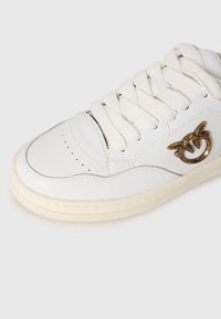 Sneaker bianco con lacci bianchi spessi, dettagli cuciti e logo in metallo dorato con due uccelli sul lato.