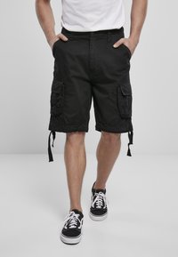 Man som bär svarta cargo-shorts och svart-vita sneakers, med händerna i fickorna, stående på en enfärgad ljus bakgrund.