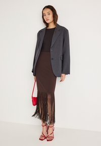 Grijze blazer, bruine rok met franjes aan de zoom, rode handtas en rode sandalen met bandjes. De stof oogt glad met een getailleerde pasvorm en een duidelijke silhouet.