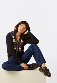 Gilet en tricot noir avec une broderie florale colorée sur le devant, associé à un jean large bleu foncé et des mocassins marron foncé.