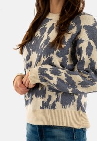 Pull en tricot avec une base crème et un motif abstrait bleu, manches et hem côtelés. Texture douce, coupe décontractée et manches longues.