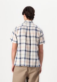 Camisa de manga curta em padrão xadrez, feita de um tecido leve. Apresenta padrões de quadrados azuis, laranja e verdes sobre um fundo creme. Gola clássica com yoke nas costas.