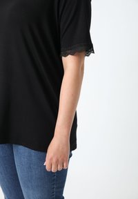 Zwart T-shirt met kanten afwerking aan de mouwen, gedragen met blauwe spijkerbroek. De stof lijkt zacht; het ontwerp is casual met een ontspannen pasvorm.