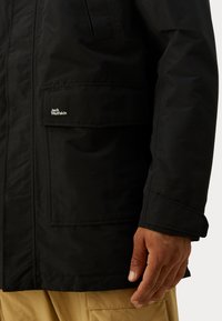 Čierna bunda so štruktúrovanou tkaninou, ktorá obsahuje náprsné vrecko. Obsahuje aj logo nášivku a má mierne oversized strih so štruktúrovanými rukávmi.