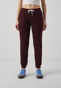 Bordeauxfarbene Sweatpants mit einem weißen Kordelzug, ausgestattet mit seitlichen Taschen und gerippten Bündchen, kombiniert mit blauen Turnschuhen. Glatte Stoffstruktur.