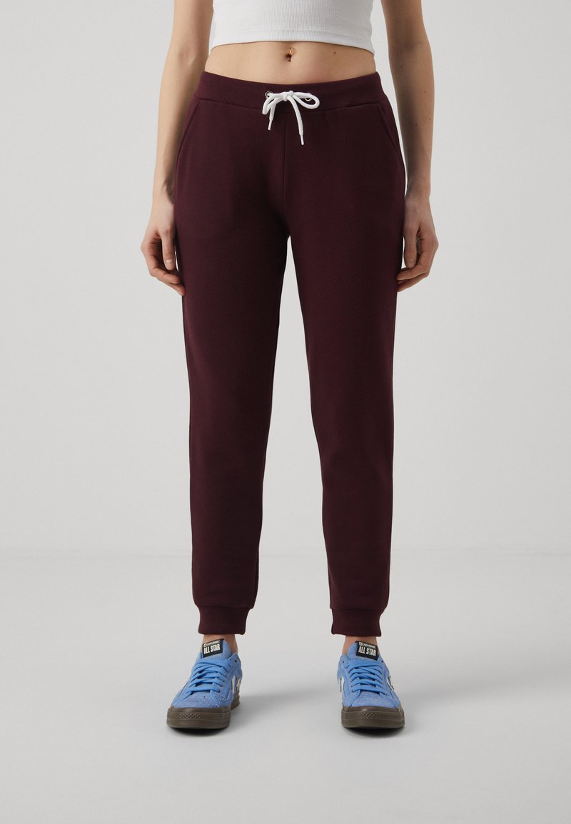Bordeauxfarbene Sweatpants mit einem weißen Kordelzug, ausgestattet mit seitlichen Taschen und gerippten Bündchen, kombiniert mit blauen Turnschuhen. Glatte Stoffstruktur.
