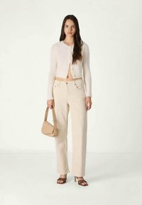 Kremni pleteni kardigan, bež hlače visokog struka i smeđe sandale s visokom petom. Drži malu bež torbicu. Jednostavna pozadina.