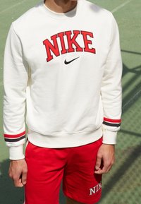 Vit sweatshirt med röd "NIKE"-text, svart logotyp och randiga muddar. Kombinerad med röda shorts med "NIKE"-broderi.