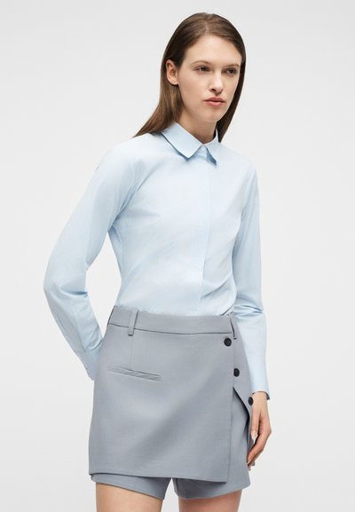 Γυναίκα που φορά ένα γαλάζιο πουκάμισο με γιακά, μπαλωμένο σε μια γκρι φούστα τύπου περιτύλιξης με κουμπιά, που στέκεται μπροστά σε απλό λευκό φόντο.