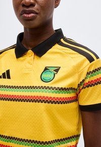 Άτομο που φοράει κίτρινη φανέλα της Ομοσπονδίας Ποδοσφαίρου της Τζαμάικα με μαύρο γιακά και πολύχρωμες ρίγες με σχέδιο στο στήθος και τα μανίκια.