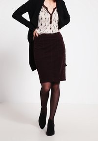 Schwarzer langer Cardigan über einer gemusterten Bluse, kombiniert mit einem dunkelbraunen Cordrock und schwarzen Stiefeletten. Einfaches Design, strukturierter Stoff.