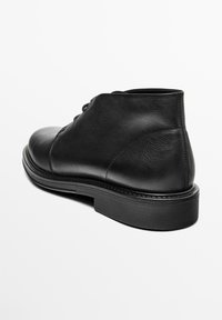 Bottine en cuir noir avec fermeture à lacets à l'avant, texture lisse et semelle en caoutchouc épaisse, vue de l'arrière sur fond blanc.