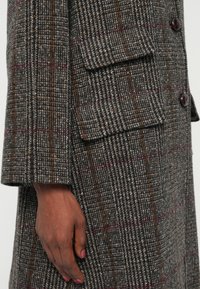 Main avec du vernis à ongles rose reposant à côté d'une poche de manteau en laine à carreaux marron et gris avec des boutons visibles sur la manche et à l'avant.